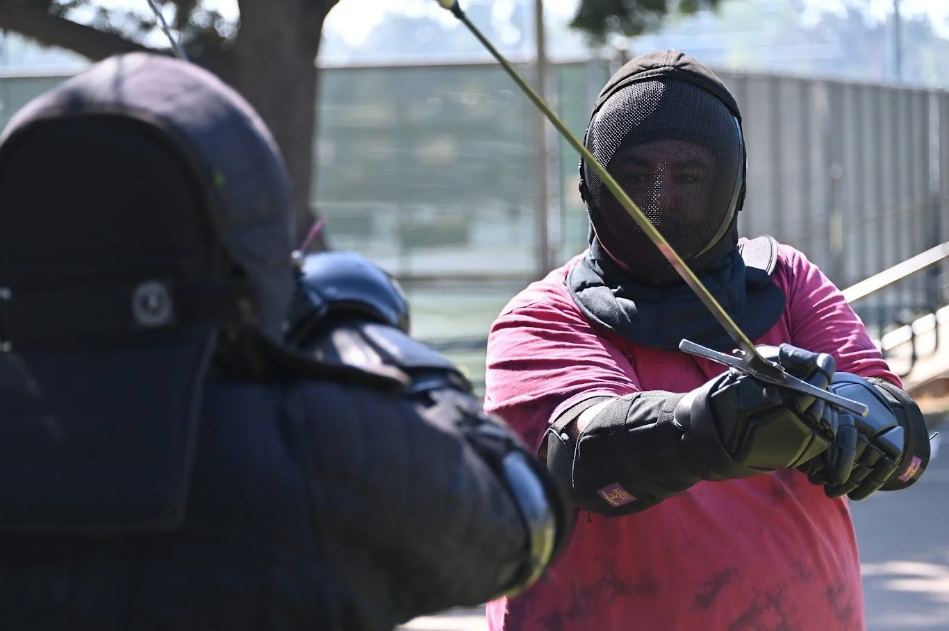 Person wearing fencing gear, including a mask, gloves, and protective clothing, in a fencing stance with a fencing foil, outdoors near a fence and trees.