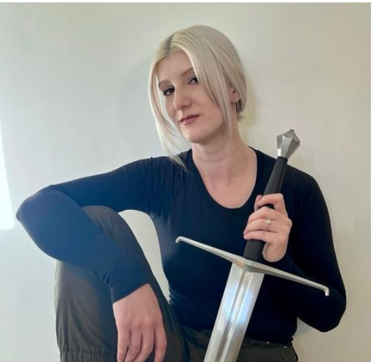 A woman with blonde hair wearing a black shirt, holding a sword vertically in front of her, sitting on a surface with one arm resting on her knee, in front of a plain light-colored wall.