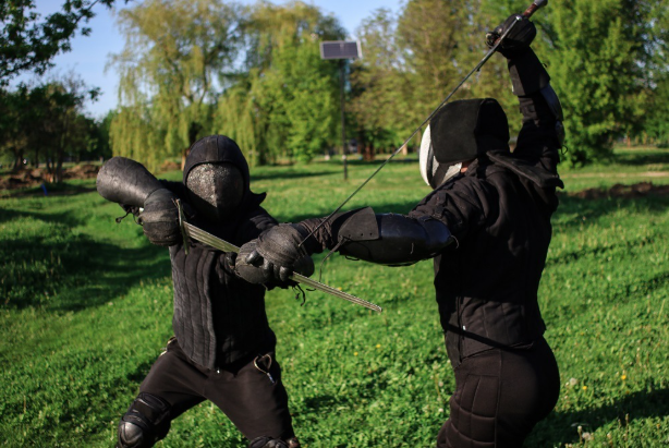 Two individuals practicing fencing outdoors in a grassy park area with trees, wearing black protective gear and masks, engaged in a fencing duel.