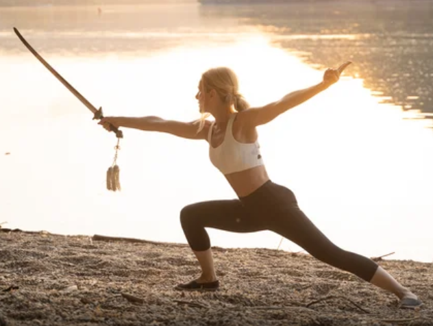 A woman practicing yoga outdoors by a lake at sunset, in warrior pose while holding a sword.