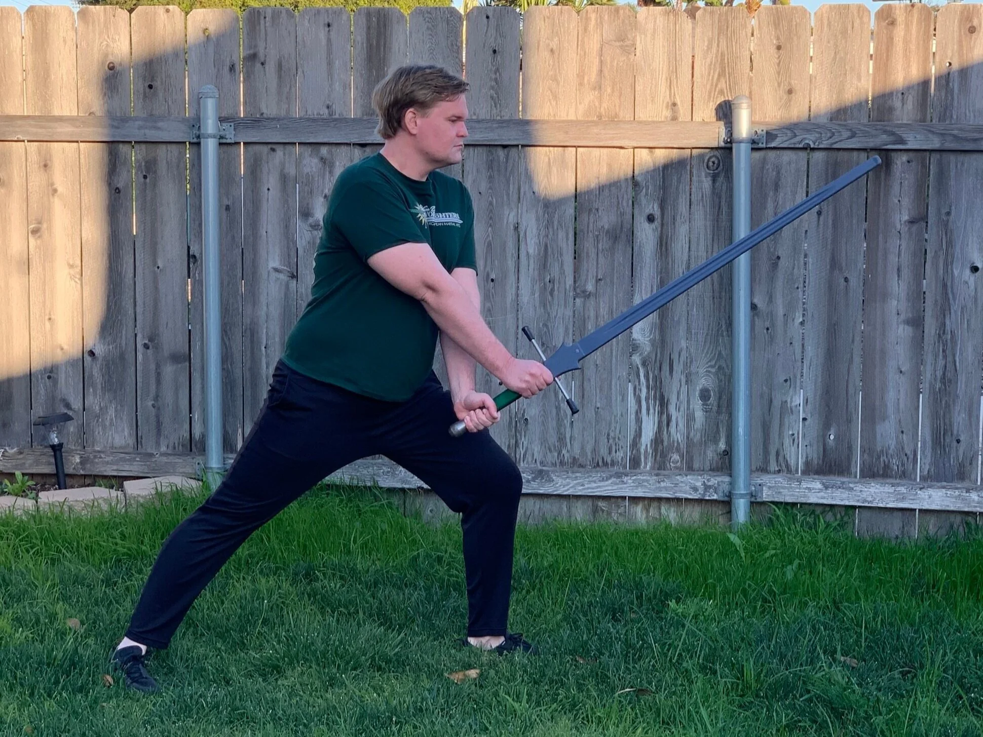 A man outdoors in a backyard holding a sword with both hands, in a broad stance with one leg forward and one leg back, facing to the right. He has light brown hair and is wearing a green t-shirt and black pants, with a wooden fence in the background and green grass on the ground.